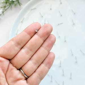 May include: A close-up of a small, silver stud earring with a clear, round stone. The earring is held between fingers, with other earrings scattered on a white plate. The background includes white flowers and a white plate.