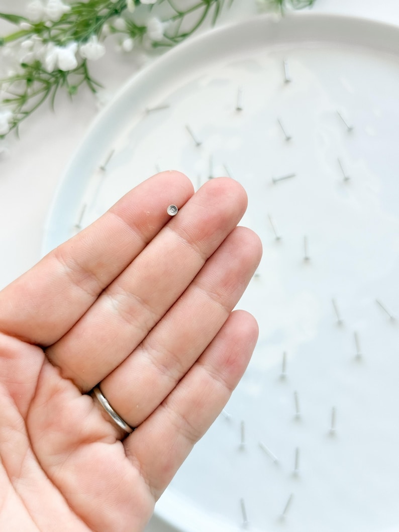 May include: A close-up of a small, silver stud earring with a clear, round stone. The earring is held between fingers, with other earrings scattered on a white plate. The background includes white flowers and a white plate.