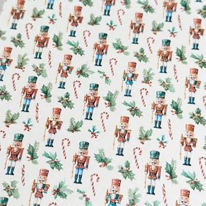May include: A repeating pattern of watercolour Christmas imagery. The design features toy nutcrackers in red, blue, and gold uniforms, candy canes, and sprigs of holly on a cream background.