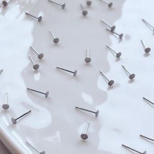 May include: A scattering of small, silver stud earrings on a white surface. Each earring features a flat, circular front and a slender post. The earrings are evenly distributed, presenting a minimalist design.