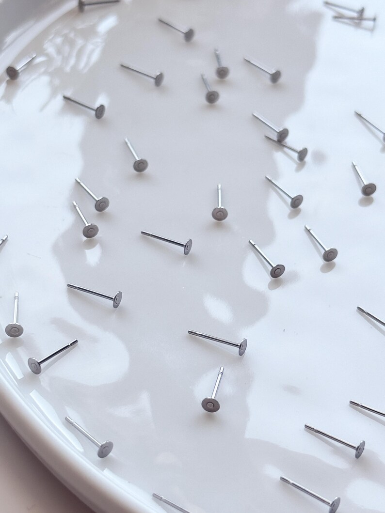 May include: A scattering of small, silver stud earrings on a white surface. Each earring features a flat, circular front and a slender post. The earrings are evenly distributed, presenting a minimalist design.