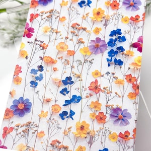 Op de afbeelding: Een rechthoekige kaart met een levendig bloemmotief. De kaart is versierd met een reeks kleurrijke geperste bloemen, waaronder oranje, gele, blauwe en paarse bloemen, tegen een witte achtergrond. Delicate stelen en bladeren dragen bij aan de natuurlijke esthetiek.
