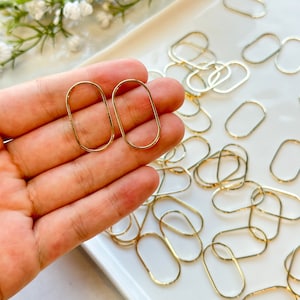 May include: Two gold oval-shaped metal rings, held in a hand, with a pile of similar rings in the background.