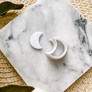 Puede incluir: Dos cortadores de galletas en forma de luna de cerámica blanca sobre una superficie de mármol blanco. El cortador más grande tiene forma de media luna, y el más pequeño tiene forma de media luna.