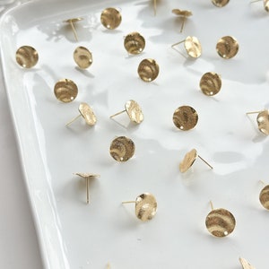 May include: A collection of gold-toned metal earring findings with a textured, hammered finish. The findings are round and have a small hole in the center for attaching earring posts.