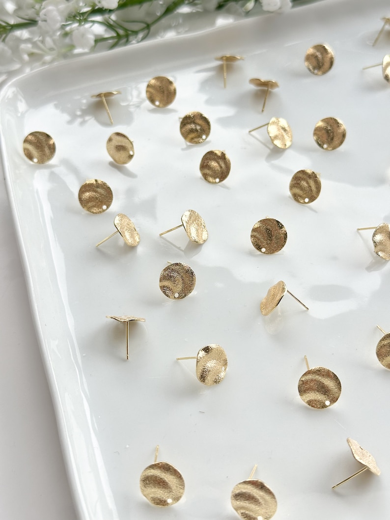 May include: A collection of gold-toned metal earring findings with a textured, hammered finish. The findings are round and have a small hole in the center for attaching earring posts.