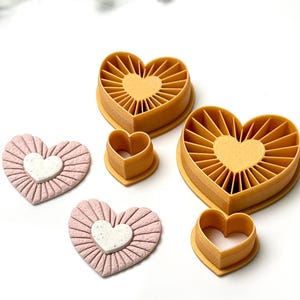 Puede incluir: Una colección de cortadores de galletas en forma de corazón y corazones decorativos. Los cortadores son de color amarillo cálido, con un diseño de rayos de sol. Los corazones decorativos son rosas con un centro blanco, mostrando una superficie texturizada.