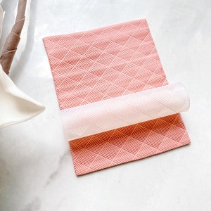 May include: A pink silicone rolling mat with a geometric pattern is being rolled with a clear silicone roller.