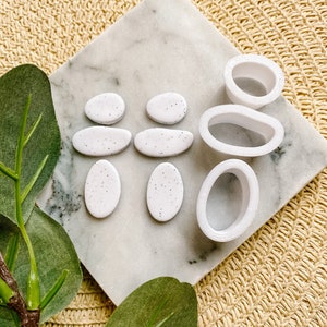 Puede incluir: Pendientes de arcilla polimérica ovalados de color blanco moteado y tres moldes de silicona blancos para hacer pendientes de arcilla polimérica.