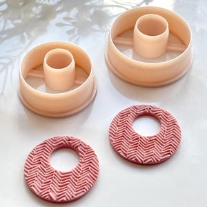 Könnte beinhalten: Zwei hellrosa Polymer Clay Ausstecher und zwei passende rosa Tonohrringe mit einem strukturierten Fischgrätenmuster. Die Ausstecher sind kreisförmig mit einem zentralen Zylinder. Die Ohrringe haben ein Loch in der Mitte zum Aufhängen.