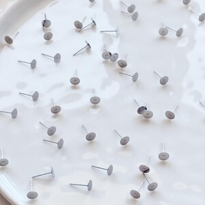 May include: A scattering of silver earring posts with flat, circular pads. The posts are arranged on a white, slightly curved surface. These small earring posts have a simple, minimalist design, suitable for jewellery making.