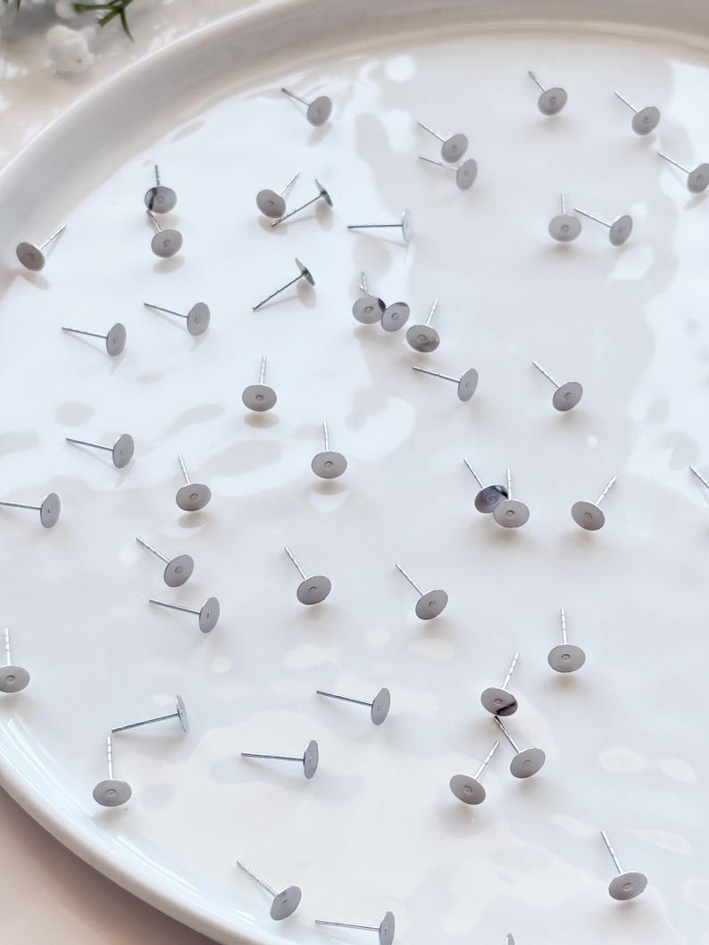 May include: A scattering of silver earring posts with flat, circular pads. The posts are arranged on a white, slightly curved surface. These small earring posts have a simple, minimalist design, suitable for jewellery making.