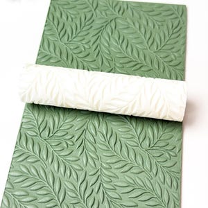 Op de afbeelding: Een witte deegroller met een bladerenpatroon ligt op een groene, getextureerde plaat met een bijpassend bladerenontwerp. De deegroller en plaat zijn waarschijnlijk voor knutselen of bakken.
