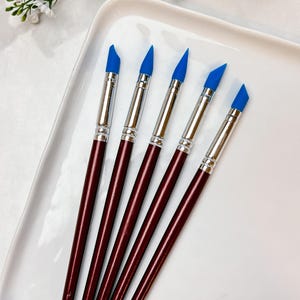 May include: Five art tools with burgundy handles and silver ferrules, each topped with a blue silicone tip. The tools are arranged on a white rectangular tray, with floral accents in the background. These tools are ideal for crafting and art projects.