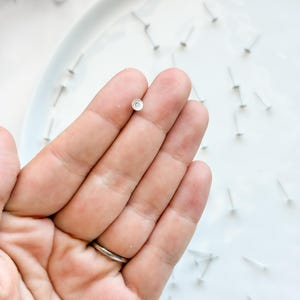 May include: A single, small silver earring stud with a circular design is held in a hand. The earring is on a white plate, surrounded by other earring posts. The hand has a silver ring on the ring finger.