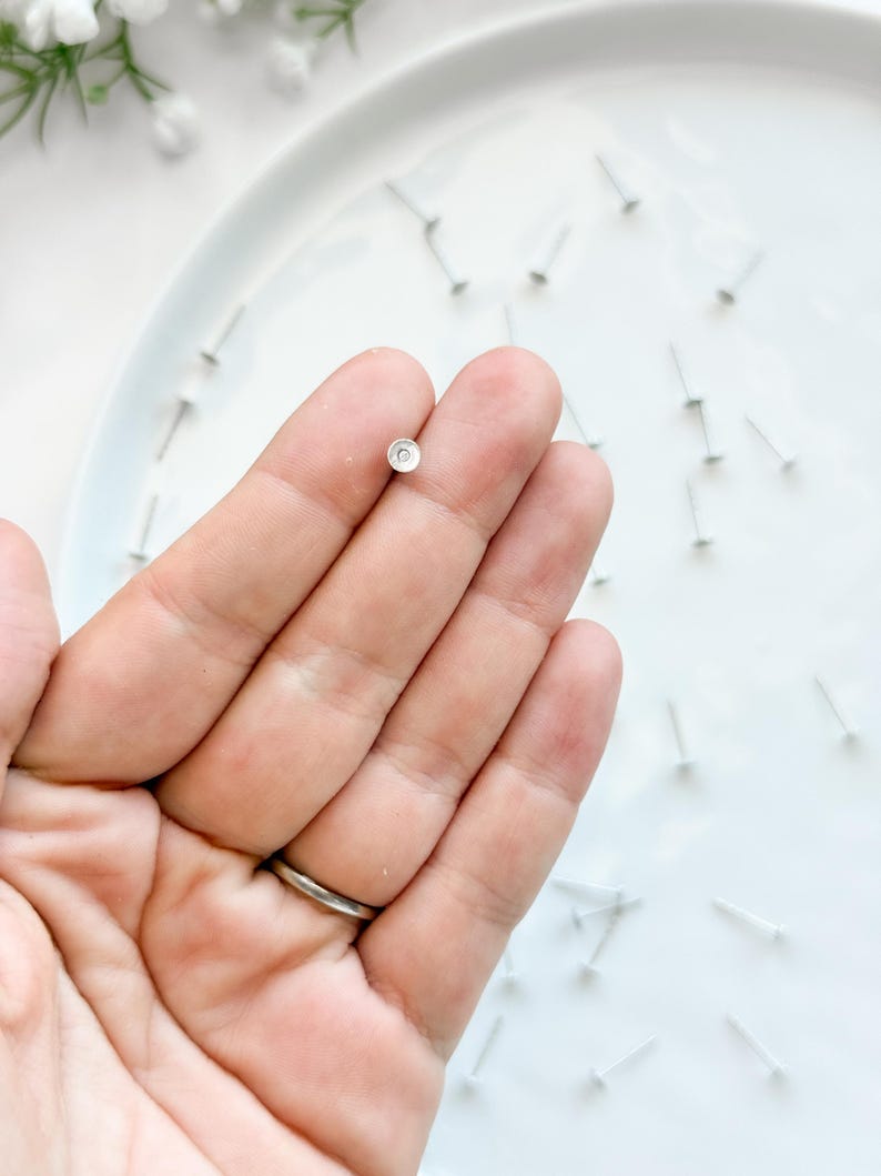 May include: A single, small silver earring stud with a circular design is held in a hand. The earring is on a white plate, surrounded by other earring posts. The hand has a silver ring on the ring finger.