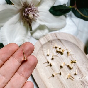 May include: A close-up of a gold earring stud with a small round back. The earring is being held by a hand. A wooden tray with a pile of similar earring studs is in the background.