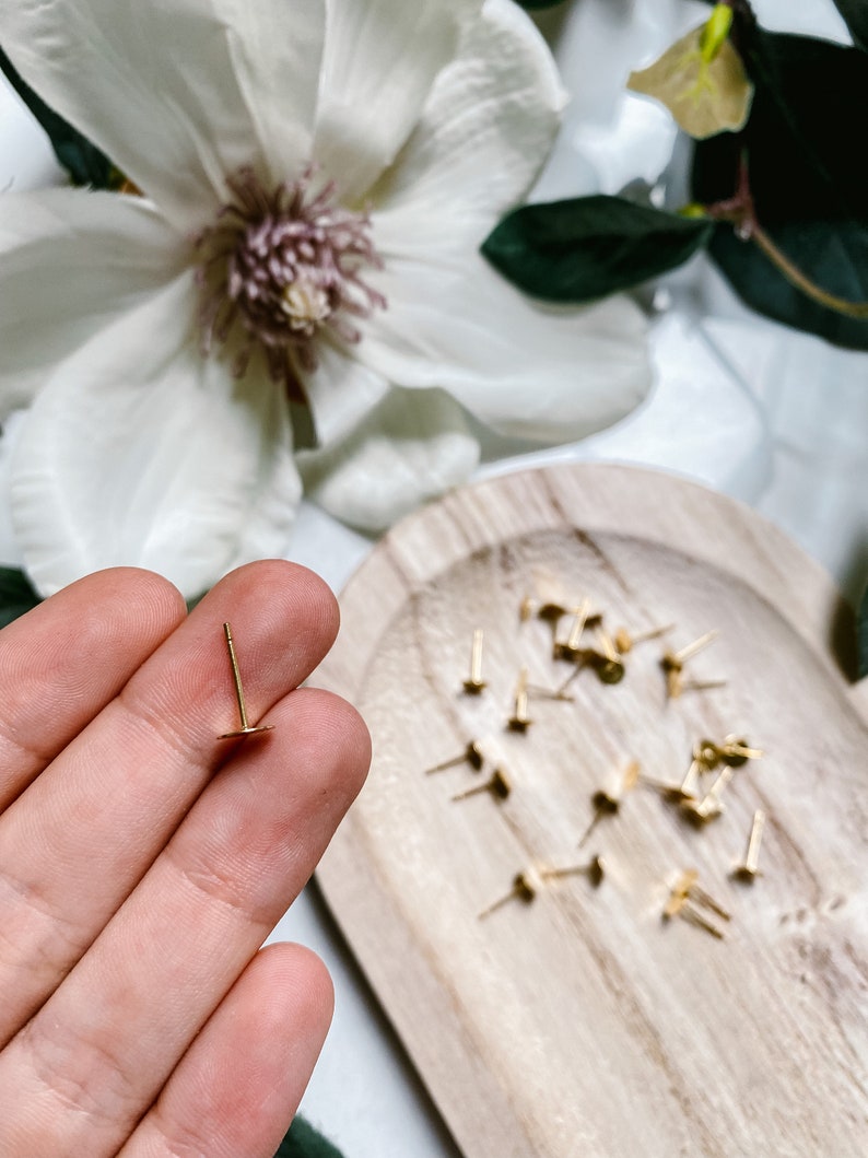 May include: A close-up of a gold earring stud with a small round back. The earring is being held by a hand. A wooden tray with a pile of similar earring studs is in the background.
