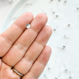 May include: A hand holding a small, silver-coloured jewellery finding. The finding has a flat, circular top and a thin post. Several similar findings are scattered on a white surface.