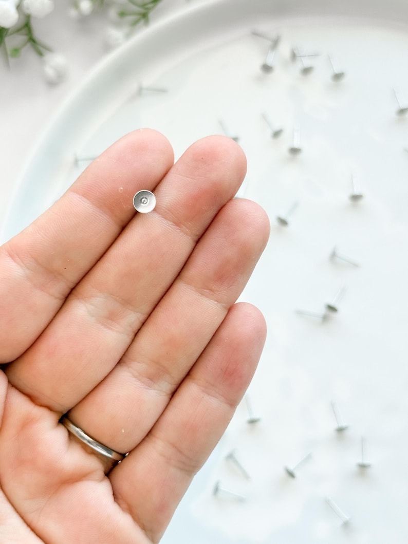 May include: A hand holding a small, silver-coloured jewellery finding. The finding has a flat, circular top and a thin post. Several similar findings are scattered on a white surface.