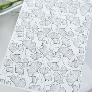 May include: A black and white illustration of a repeating pattern of ginkgo leaves. The leaves are drawn with fine lines and are arranged in a random pattern.