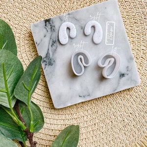 Könnte beinhalten: Zwei Paare weiße Polymer Clay Ohrringe mit gesprenkelter Textur. Die Ohrringe sind in Form einer geschwungenen Linie. Die Ohrringe werden auf einer weißen Marmorfliese mit einem Zollmaßstab angezeigt. Die Fliese befindet sich auf einer gewebten Strohmatte.