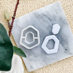 May include: Two white geometric cookie cutters on a marble surface. The larger cutter is a hexagon with a smaller hexagon cut out of the centre. The smaller cutter is a smaller hexagon.