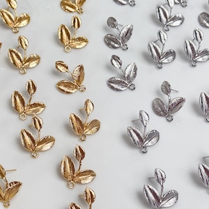 Puede incluir: Un surtido de pendientes de botón con forma de hoja en tonos dorados y plateados. Los pendientes presentan un diseño detallado y texturizado con tres hojas y un pequeño lazo en la base. Los pendientes están dispuestos sobre una superficie blanca.
