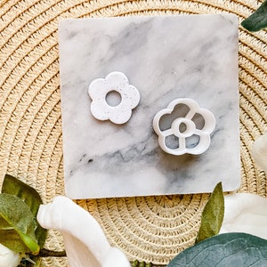 Op de afbeelding: Twee witte plastic bloemvormige koekjesvormen op een marmeren oppervlak. De grotere vorm heeft een effen ontwerp, terwijl de kleinere vorm een ​​meer ingewikkeld ontwerp heeft met uitsnijdingen.
