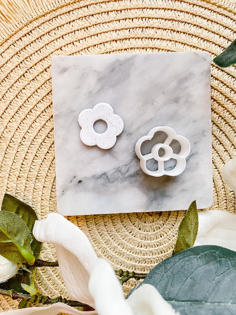 Op de afbeelding: Twee witte plastic bloemvormige koekjesvormen op een marmeren oppervlak. De grotere vorm heeft een effen ontwerp, terwijl de kleinere vorm een ​​meer ingewikkeld ontwerp heeft met uitsnijdingen.