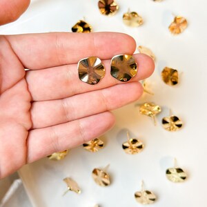 May include: A close-up of a hand holding two gold-toned, wavy, round earring blanks. The blanks are on a white surface with other similar blanks scattered around.