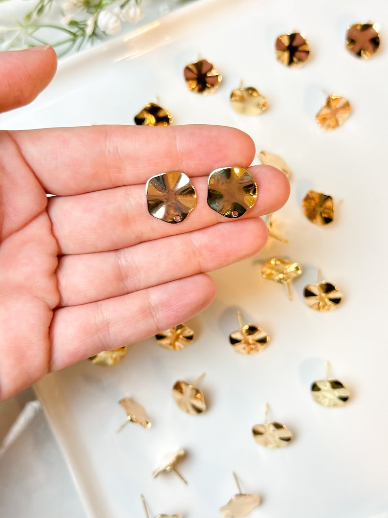 May include: A close-up of a hand holding two gold-toned, wavy, round earring blanks. The blanks are on a white surface with other similar blanks scattered around.