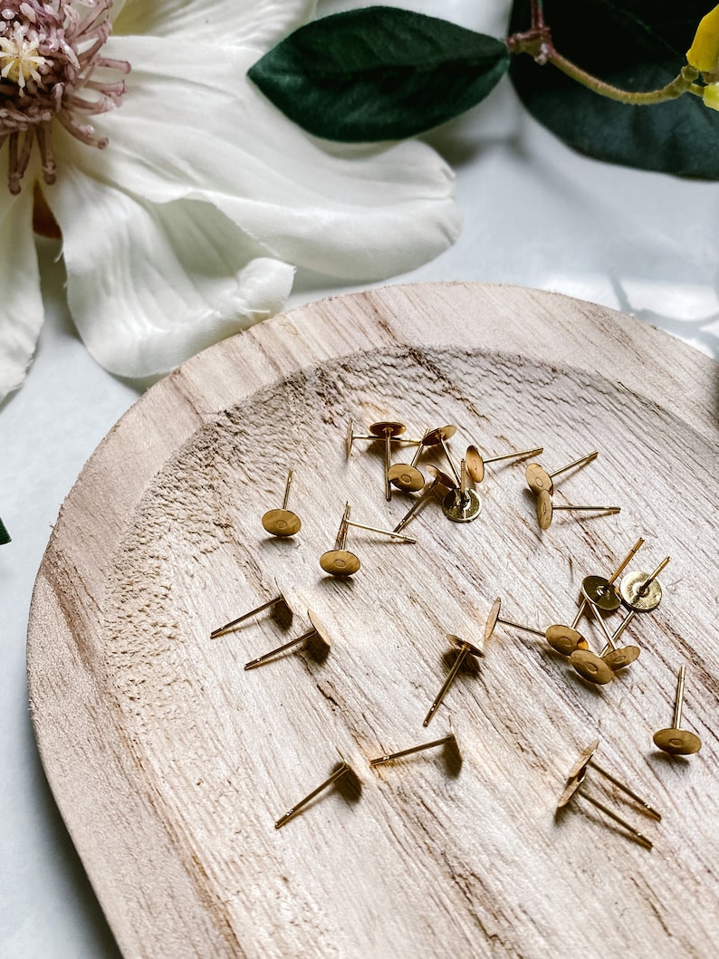 May include: A close-up of a wooden plate with a scattering of gold earring posts and backs. The earring posts are made of metal and have a small, round head. The earring backs are also made of metal and have a flat, round shape.