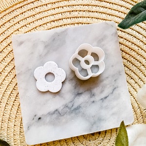 Op de afbeelding: Twee witte plastic bloemvormige koekjesvormen op een marmeren oppervlak. De grotere vorm heeft een meer gedetailleerd ontwerp met vijf bloemblaadjes en een gat in het midden. De kleinere vorm heeft een eenvoudiger ontwerp met vier bloemblaadjes en een gat in het midden.