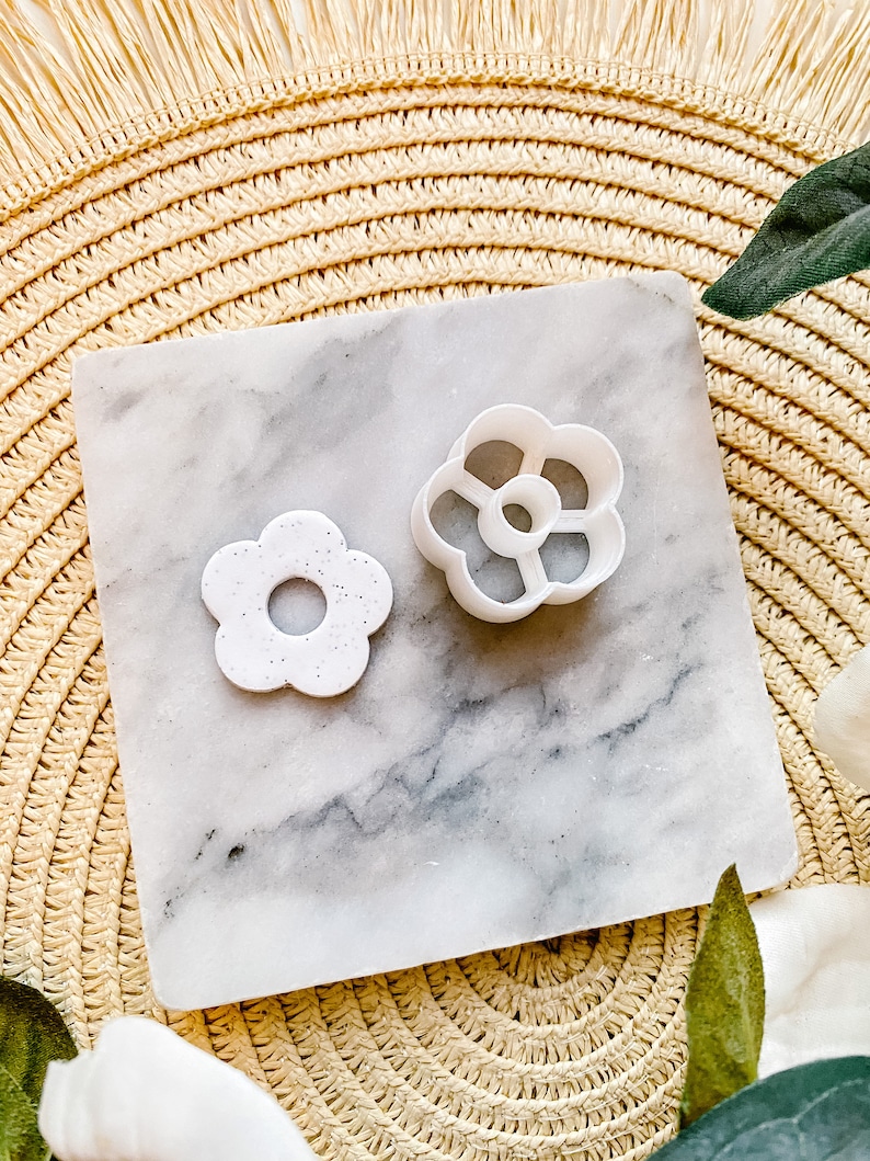 Op de afbeelding: Twee witte plastic bloemvormige koekjesvormen op een marmeren oppervlak. De grotere vorm heeft een meer gedetailleerd ontwerp met vijf bloemblaadjes en een gat in het midden. De kleinere vorm heeft een eenvoudiger ontwerp met vier bloemblaadjes en een gat in het midden.