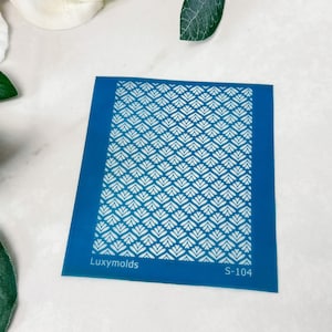 Può includere: Uno stencil di plastica blu con un motivo geometrico bianco. Lo stencil ha il testo "Luxymolds" e "S-104" stampato su di esso.