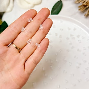 Könnte beinhalten: Eine Hand hält mehrere kleine, transparente Ohrringverschlüsse. Die Ohrringverschlüsse bestehen aus transparentem Material und sind auf einem weißen Teller verstreut. Ein goldener Ring ist an der Hand zu sehen.