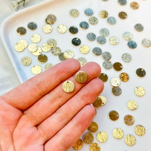 Peut inclure: Gros plan sur une main tenant deux petits charmes ronds en métal doré avec une surface texturée et un petit trou en haut. Les charmes sont entourés d'une surface blanche avec de nombreux charmes similaires éparpillés autour.