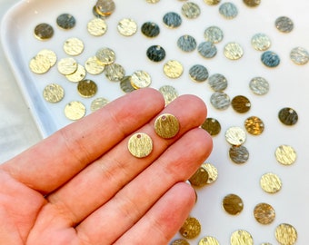 May include: A close-up of a hand holding two small, gold, round metal charms with a textured surface and a small hole at the top. The charms are surrounded by a white surface with many similar charms scattered around.