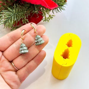 以下が含まれることがあります： 緑と白のクリスマスツリーのチャームが付いた、金色のフープイヤリングのペア。クリスマスツリーのデザインが施された黄色のシリコンモールドも写っています。