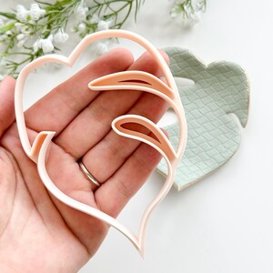 May include: A pink plastic cookie cutter shaped like a monstera leaf. The cutter is held in a hand and is being shown next to a green and white patterned surface.