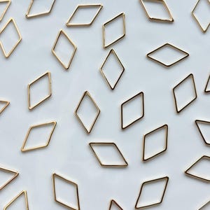 Puede incluir: Vista aérea de numerosos marcos metálicos dorados en forma de rombo dispersos sobre una superficie blanca. Los marcos son delgados y tienen un acabado pulido, creando un patrón geométrico simple. Los rombos son todos del mismo tamaño.