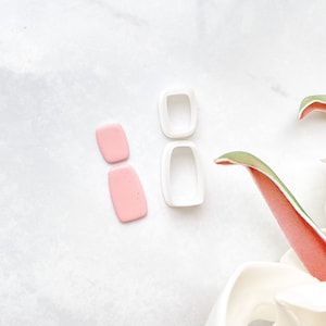 May include: Two pink rectangular polymer clay pieces and two white rectangular cutters are displayed on a white surface. The clay pieces are a soft pink colour, and the cutters are white. The image has a clean, minimalist aesthetic.