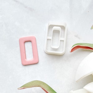 Op de afbeelding: Twee koekjesvormen, een witte en een roze, in de vorm van een rechthoek met een gat in het midden. De vormen liggen op een wit marmeren oppervlak.