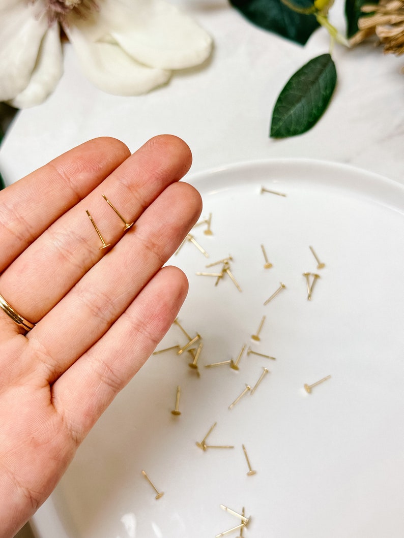 May include: A close-up of a hand holding two gold earring posts. A white plate with a scattering of gold earring posts is in the background.