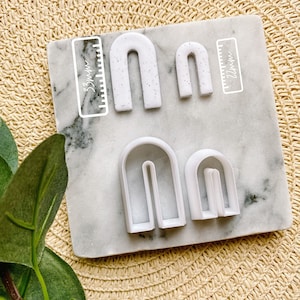 May include: Set of four white arch-shaped cookie cutters on a white marble surface. The cutters are 3.8 cm and 2.2 cm in size.