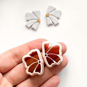 May include: Two sets of flower-shaped polymer clay cutters. The top set is gray with gold centers. The bottom set is a reddish-brown color with a white outline. The cutters are for crafting.