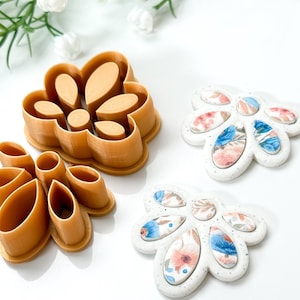 Puede incluir: Dos cortadores de galletas con forma de flor en un tono marrón cálido se encuentran junto a dos diseños florales terminados. Los cortadores son de plástico. Las flores terminadas tienen bases blancas con coloridos patrones florales en azul, rosa y melocotón.