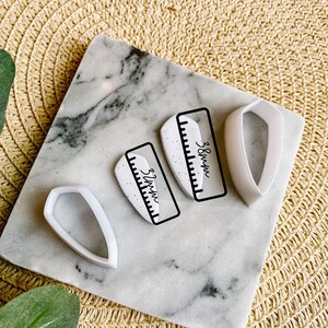 Op de afbeelding: Twee witte siliconen mallen voor het maken van harssieraden. De mallen zijn in de vorm van een traan en een rechthoek. De mallen liggen op een wit marmeren oppervlak. De rechthoekige mal heeft een liniaal erop gedrukt met de afmetingen 5,5 cm en 9 cm.