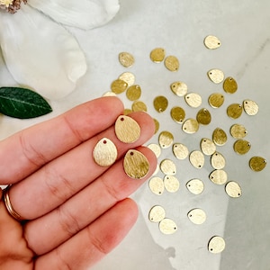 Może przedstawiać: Kolekcja małych, złotych metalowych zawieszek w kształcie łezki o teksturowanej powierzchni. Każda zawieszka ma mały otwór u góry do nawlekania. Zawieszki są rozrzucone na jasnej powierzchni, niektóre trzymane w dłoni.